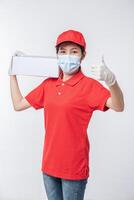 imagen de un joven repartidor consciente con gorra roja en blanco, camiseta uniforme, guantes de máscara facial de pie con una caja de cartón blanca vacía aislada en un estudio de fondo gris claro foto