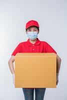 imagen de un joven repartidor consciente con gorra roja en blanco camiseta uniforme guantes de máscara facial de pie con una caja de cartón marrón vacía aislada en un estudio de fondo gris claro foto