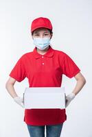 Image of a conscious young delivery man in red cap blank t-shirt uniform face mask gloves standing with empty white cardboard box isolated on light gray background studio photo