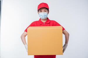 Image of a conscious young delivery man in red cap blank t-shirt uniform face mask gloves standing with empty brown cardboard box isolated on light gray background studio photo