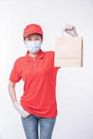 Image of a happy young delivery man in red cap blank t-shirt uniform face mask gloves standing with empty brown craft paper packet isolated on light gray background studio photo