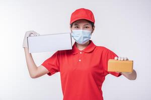 Image of a conscious young delivery man in red cap blank t-shirt uniform face mask gloves standing with empty white cardboard box isolated on light gray background studio photo