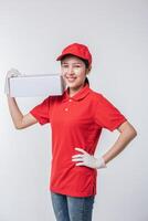 imagen de un joven repartidor con uniforme de camiseta en blanco con gorra roja de pie con una caja de cartón blanca vacía aislada en un estudio de fondo gris claro foto