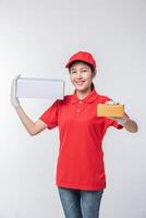 imagen de un joven repartidor con uniforme de camiseta en blanco con gorra roja de pie con una caja de cartón blanca vacía aislada en un estudio de fondo gris claro foto