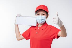 Image of a conscious young delivery man in red cap blank t-shirt uniform face mask gloves standing with empty white cardboard box isolated on light gray background studio photo