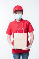 Image of a happy young delivery man in red cap blank t-shirt uniform face mask gloves standing with empty brown craft paper packet isolated on light gray background studio photo