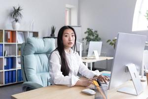 asiático mujer en el oficina. mujer de negocios, se sienta en un blanco camisa a el escritorio, trabajos a el computadora. foto
