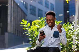 hombre afroamericano feliz con ropa informal sentado en un banco al aire libre con teléfono móvil foto