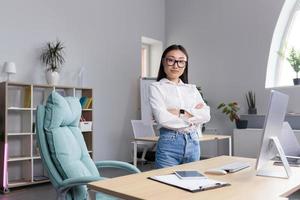 asiático mujer mujer de negocios en el oficina a el escritorio. cruzado su brazos, mira a el cámara, sonrisas foto