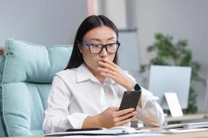 Worried young Asian business woman in glasses holds the phone in her hands, surprised, shocked. photo