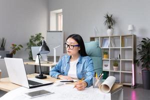 asiático diseñador trabajando en oficina, con planos, mujer a trabajo en casual ropa foto