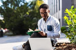 Young african american male student sitting in a city park on a bench with laptop and notebook photo