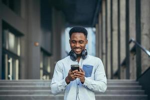 joven africano americano hombre contento con ganar mira a teléfono se regocija y sonrisas en casual ropa foto