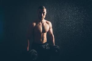 Male blond athlete lifts dumbbells with hand on biceps studio photo with raindrops