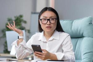 Disappointed and sad asian business woman reading bad news from phone, employee working in office photo