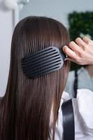 Young woman combing her long dark hair with a comb in a beauty salon. A straight healthy brunette hair that has undergone the hair straightening procedure. photo