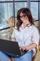 bella mujer de negocios trabajando en una laptop en una silla cerca de la ventana. mujer independiente que se conecta a internet a través de la computadora. blogger o periodista escribiendo un nuevo artículo. foto