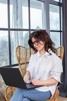 Beautiful businesswoman working on a laptop in a chair near the window. Female freelancer connecting to internet via computer. Blogger or journalist writing new article. photo