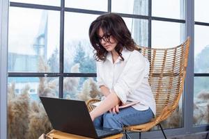 Beautiful businesswoman working on a laptop in a chair near the window. Female freelancer connecting to internet via computer. Blogger or journalist writing new article. photo