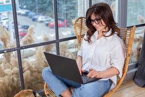 Beautiful businesswoman working on a laptop in a chair near the window. Female freelancer connecting to internet via computer. Blogger or journalist writing new article. photo