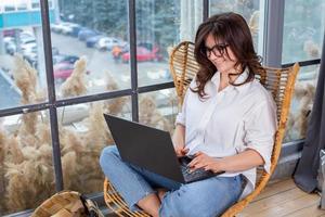 Beautiful businesswoman working on a laptop in a chair near the window. Female freelancer connecting to internet via computer. Blogger or journalist writing new article. photo