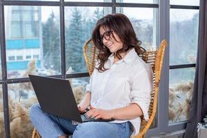 Beautiful businesswoman working on a laptop in a chair near the window. Female freelancer connecting to internet via computer. Blogger or journalist writing new article. photo
