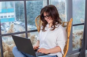Beautiful businesswoman working on a laptop in a chair near the window. Female freelancer connecting to internet via computer. Blogger or journalist writing new article. photo