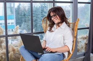 Beautiful businesswoman working on a laptop in a chair near the window. Female freelancer connecting to internet via computer. Blogger or journalist writing new article. photo