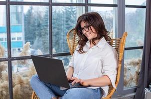 Beautiful businesswoman working on a laptop in a chair near the window. Female freelancer connecting to internet via computer. Blogger or journalist writing new article. photo