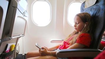 adorabile poco ragazza in viaggio di un aereo seduta vicino finestra. ragazzo ascoltando musica e invio Messaggio seduta vicino aereo finestra video