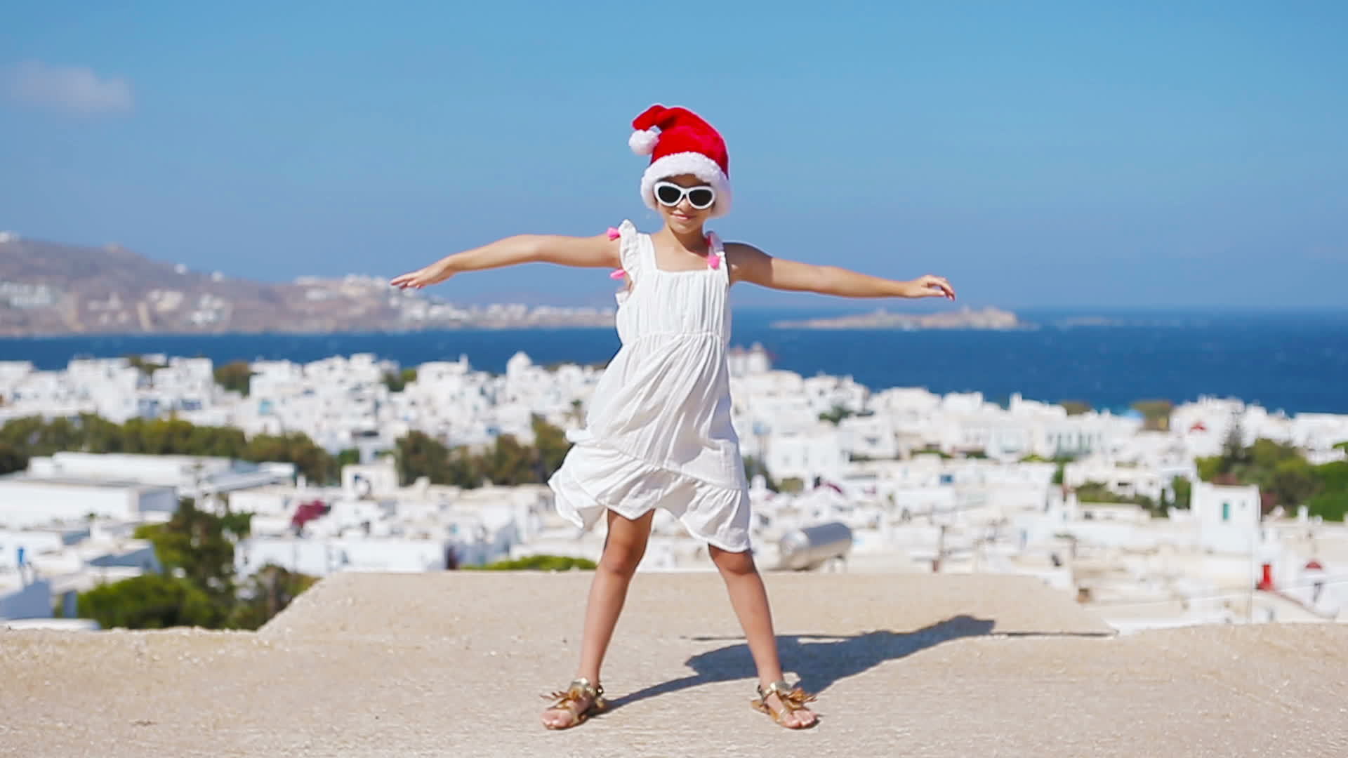 Little funny girl in red Santa hat outdoors background of Mykonos. Kid at  street of typical greek traditional village with white walls and colorful  doors on Christmas vacation in Greece 20044776 Stock