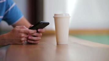 Nahaufnahme männlicher Hände, die Handy und ein Glas Kaffee im Café halten. mann, der mobiles smartphone verwendet. Junge berührt einen Bildschirm seines Smartphones. unscharfer Hintergrund, horizontal. video