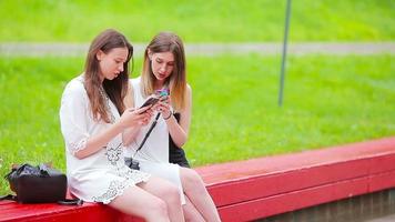 dois jovem meninas usando inteligente telefones ao ar livre. dois mulheres sentado dentro parque e enviando mensagem de Smartphone video