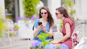 twee jong meisjes nemen selfie met slim telefoon Bij de buitenshuis cafe. twee Dames na boodschappen doen met Tassen zittend in open lucht cafe met koffie en gebruik makend van smartphone video