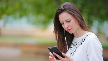 Jeune caucasien femme Envoi en cours message par sa téléphone intelligent Extérieur dans le parc. magnifique fille dans des lunettes de soleil séance sur en bois banc en utilisant téléphone intelligent video
