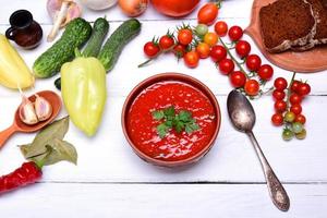 sopa gazpacho en un marrón cerámico plato en un blanco de madera fondo, parte superior vista, alrededor Fresco vegetales y hierbas para Cocinando foto