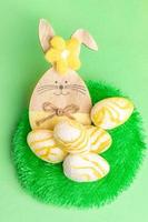 Funny wooden bunny with textured white-yellow Easter eggs on round decorative grass carpet on green. photo