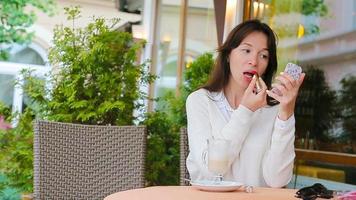 Beautiful woman painting her lips with red lipstick sitting in outdoor cafe. Young girl with delicious sweet cappuccino. video