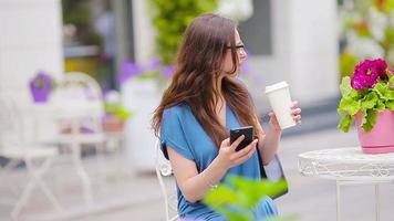 Portrait of beautiful woman sitting in outdoor cafe drinking coffee and using smartphone. Young girl looking instagram and drinking delicious sweet cappuccino. video