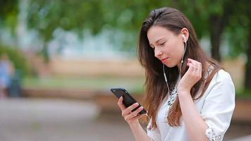 ung caucasian kvinna sändning meddelande och lyssna musik utomhus- på europeisk stad. skön flicka Sammanträde på trä- bänk använder sig av smartphone i mjuk solnedgång ljus video