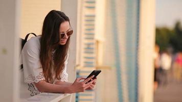 jovem caucasiano mulher enviando mensagem de dela Smartphone ao ar livre dentro a parque às pôr do sol. lindo menina dentro oculos de sol sentado em de madeira Banco usando Smartphone video