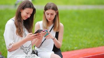zwei jung Mädchen mit Clever Telefon draußen. zwei Frauen Sitzung im Park mit Kaffee und mit Smartphone video