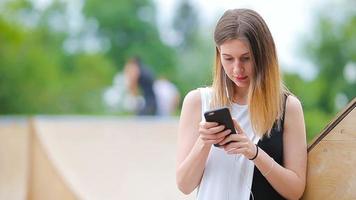 jung kaukasisch Frau Senden Botschaft und Hör mal zu Musik- draussen beim europäisch Stadt. schön Mädchen im Sonnenbrille Sitzung auf hölzern Bank mit Smartphone video