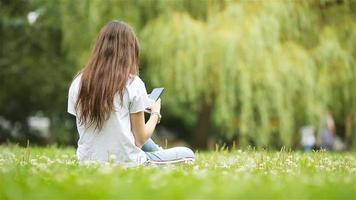 Cute woman is reading text message on mobile phone while sitting in the park. video