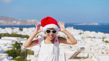 menina engraçada no chapéu vermelho de papai noel ao ar livre fundo de mykonos. garoto na rua da típica vila tradicional grega com paredes brancas e portas coloridas nas férias de natal na grécia video