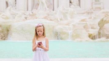 adorable pequeño niña con inteligente teléfono a calentar día al aire libre en europeo ciudad cerca famoso fontana di trevi video