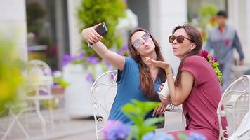 twee jong meisjes nemen selfie met slim telefoon Bij de buitenshuis cafe. twee Dames na boodschappen doen met Tassen zittend in open lucht cafe met koffie en gebruik makend van smartphone video