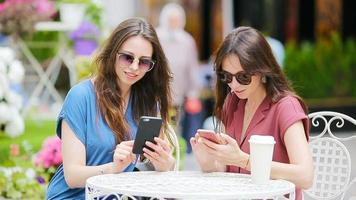 zwei jung Mädchen mit Clever Telefon beim das draußen Cafe. zwei Frauen nach Einkaufen mit Taschen Sitzung im openair Cafe mit Kaffee und mit Smartphone video