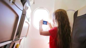 Adorable little girl traveling by an airplane sitting near window. Kid taking photos of clouds and sky sitting near aircraft window video