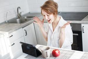 Happy surprised woman with cup of coffee or tea using laptop in the kitchen. girl with a tablet raised her hands up photo